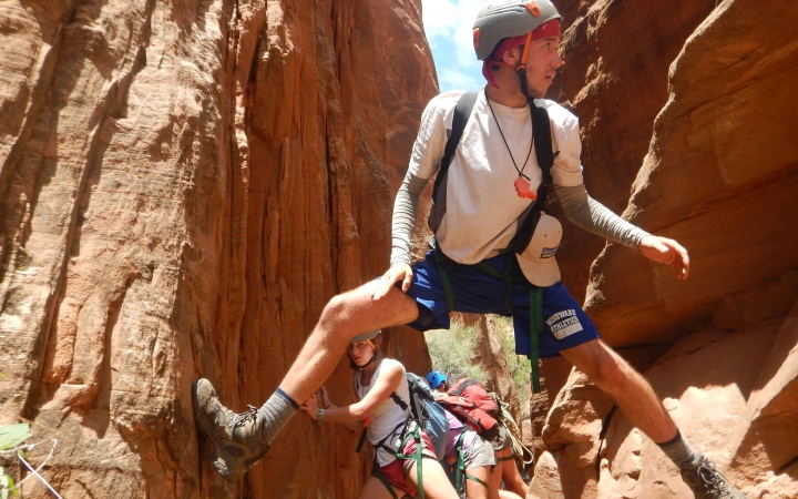 A person wearing safety gear braces their feet against the walls of a narrow canyon. Others are behind them.